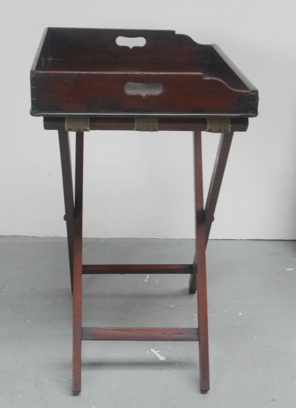 A dark mahogany butlers tray with integrated hand cutout handles resting on a matching folding x-frame stand. View VII