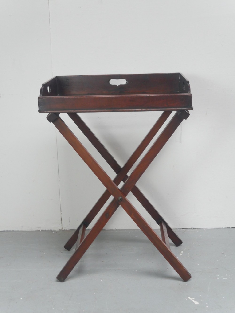 A dark mahogany butlers tray with integrated hand cutout handles resting on a matching folding x-frame stand. View XI