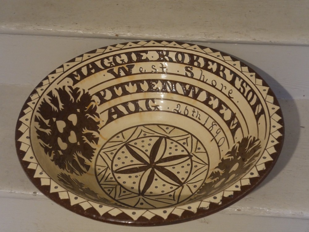 A circular glazed earthenware dairy bowl featuring dark brown slip decorations, geometric borders, central star motifs, and inscribed script lettering on a cream ground.