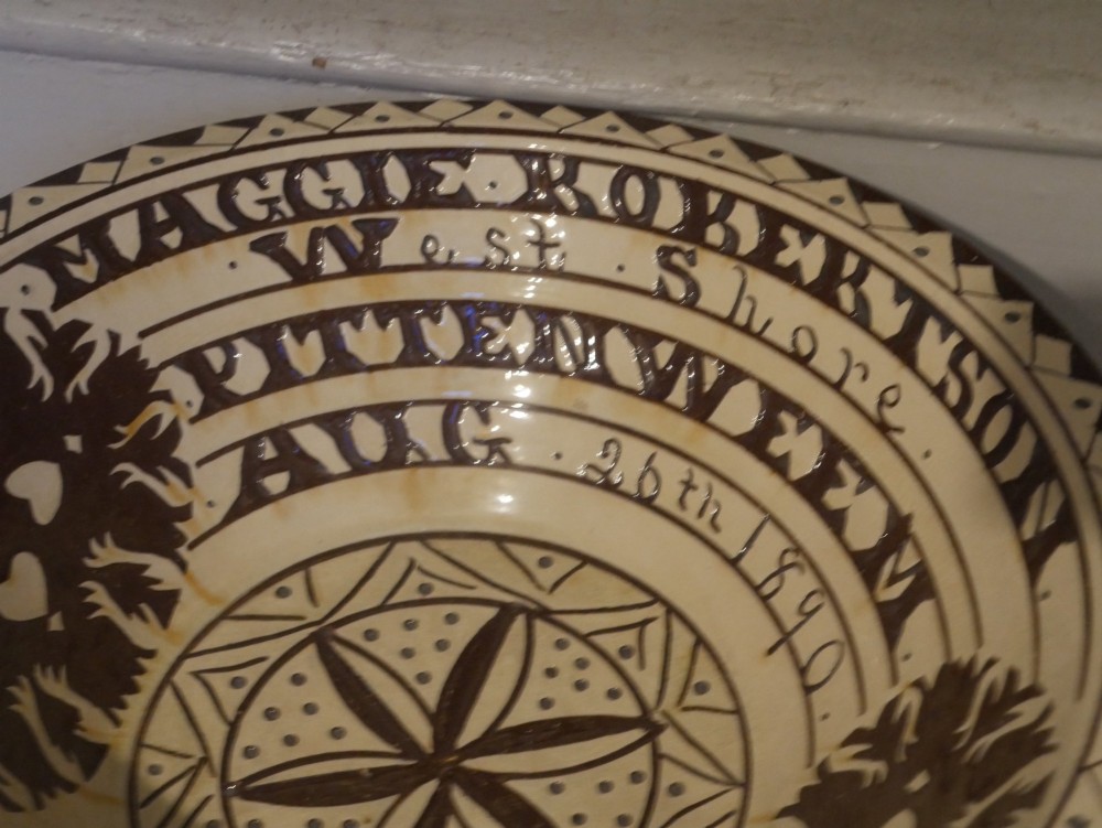 A circular glazed earthenware dairy bowl featuring dark brown slip decorations, geometric borders, central star motifs, and inscribed script lettering on a cream ground. View II