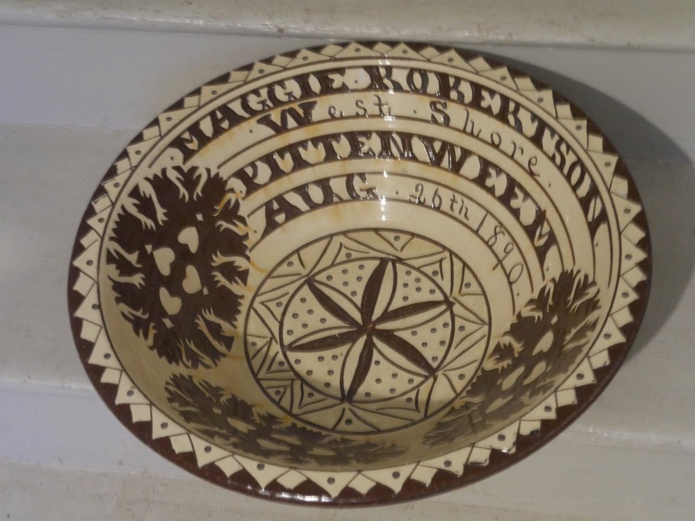 A circular glazed earthenware dairy bowl featuring dark brown slip decorations, geometric borders, central star motifs, and inscribed script lettering on a cream ground. View IV
