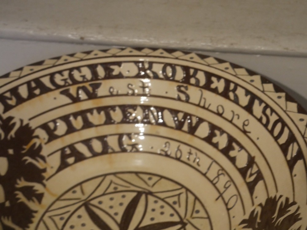 A circular glazed earthenware dairy bowl featuring dark brown slip decorations, geometric borders, central star motifs, and inscribed script lettering on a cream ground. View V