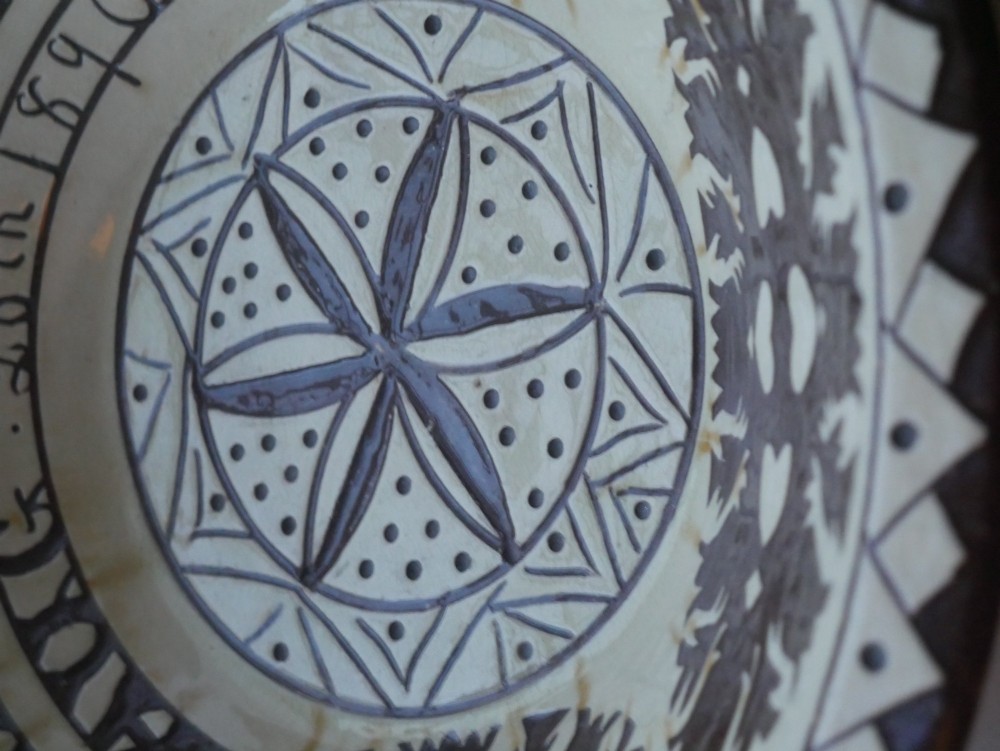 A circular glazed earthenware dairy bowl featuring dark brown slip decorations, geometric borders, central star motifs, and inscribed script lettering on a cream ground. View VI