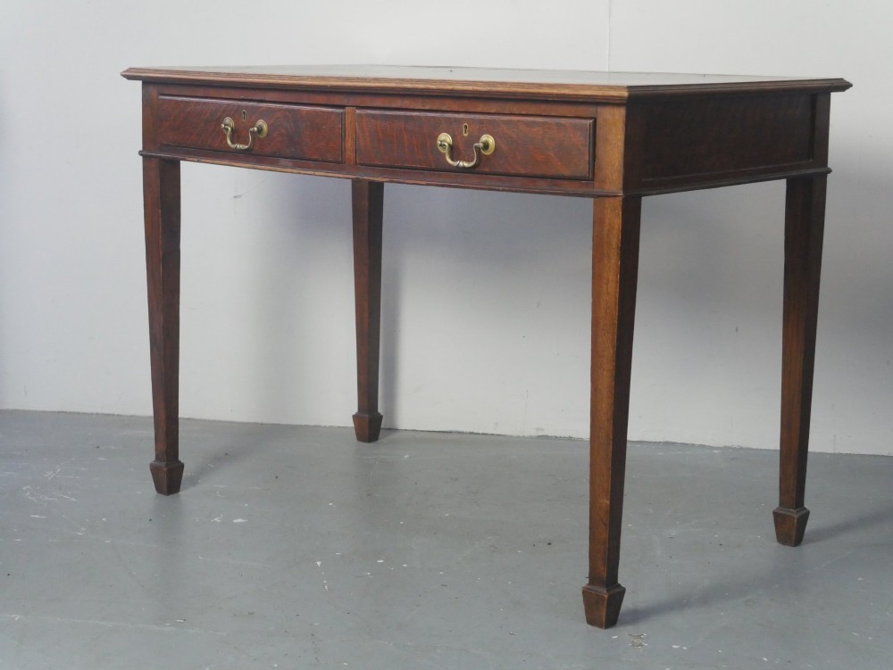 A warm brown oak writing desk with two drawer fronts featuring brass drop handles, resting on four square tapered legs.