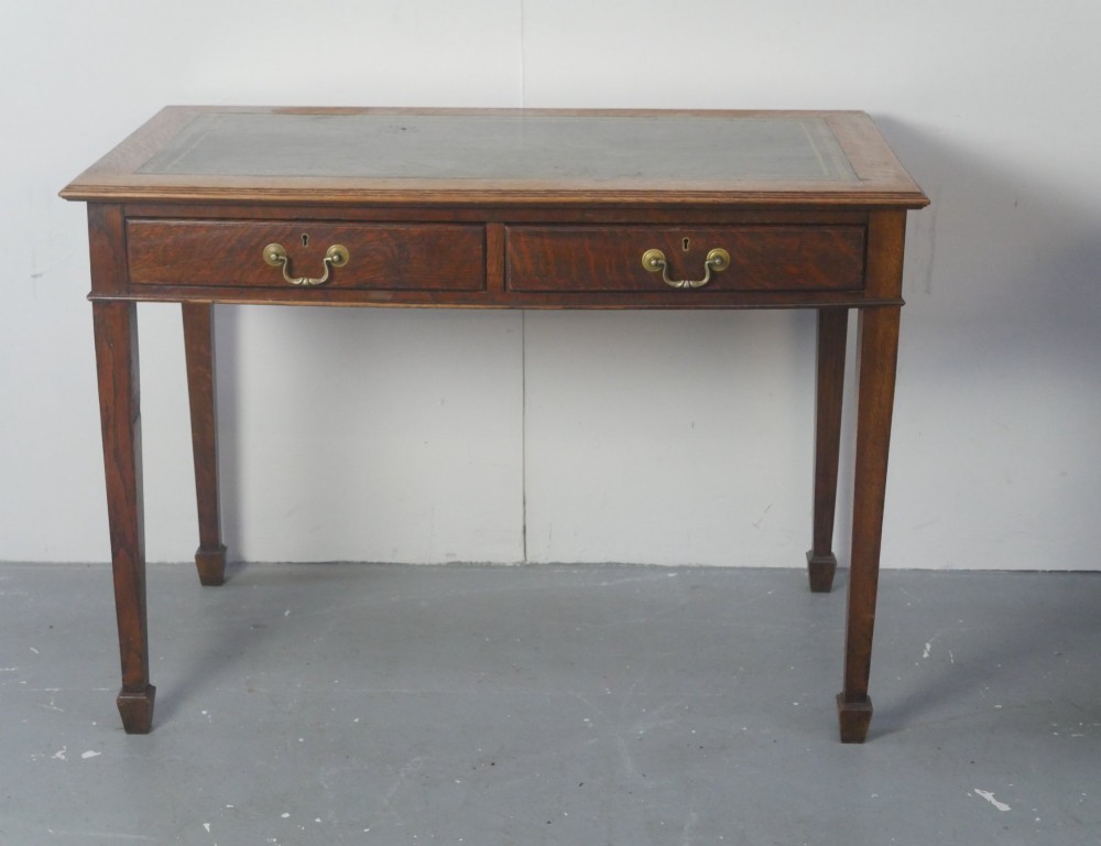A warm brown oak writing desk with two drawer fronts featuring brass drop handles, resting on four square tapered legs. View III