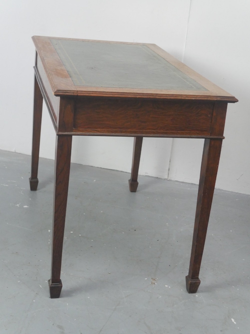 A warm brown oak writing desk with two drawer fronts featuring brass drop handles, resting on four square tapered legs. View X