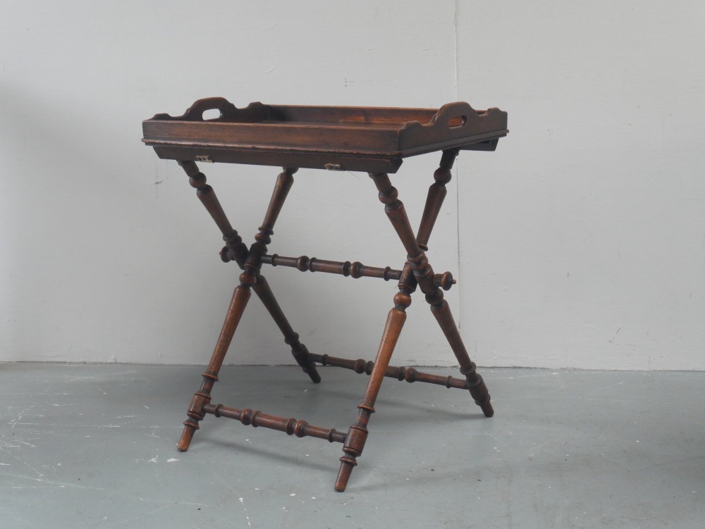 A dark oak butler tray with gallery edges and cut-out handles resting atop a matching wooden folding X-frame stand with turned legs.