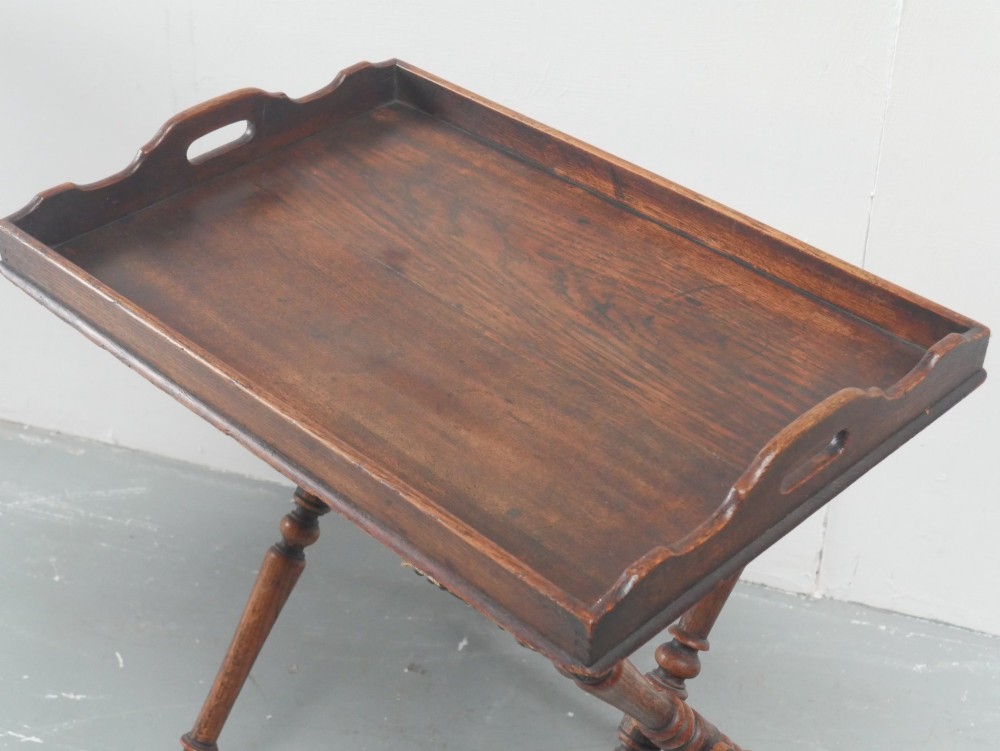 A dark oak butler tray with gallery edges and cut-out handles resting atop a matching wooden folding X-frame stand with turned legs. View II