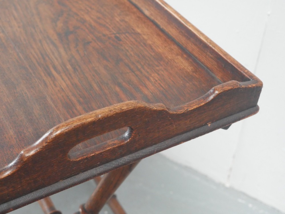 A dark oak butler tray with gallery edges and cut-out handles resting atop a matching wooden folding X-frame stand with turned legs. View III