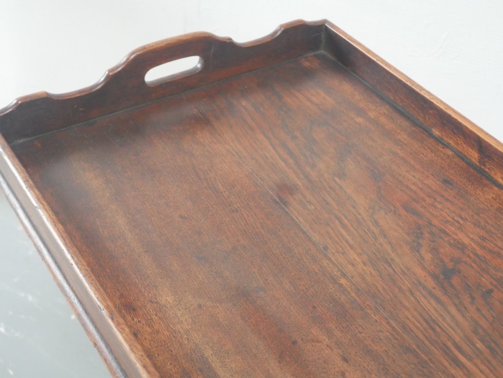 A dark oak butler tray with gallery edges and cut-out handles resting atop a matching wooden folding X-frame stand with turned legs. View VI
