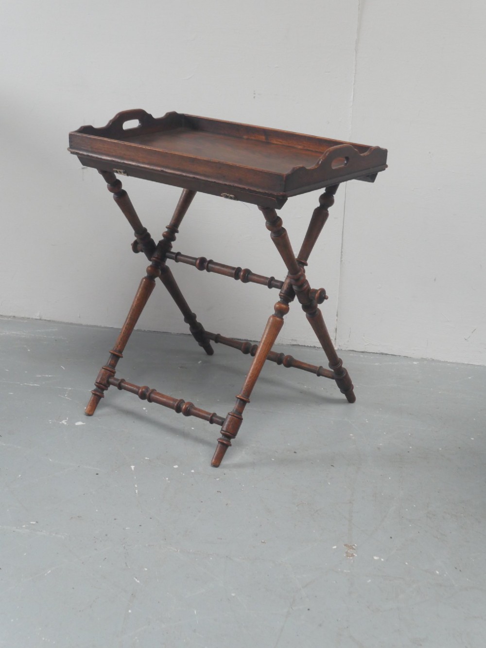 A dark oak butler tray with gallery edges and cut-out handles resting atop a matching wooden folding X-frame stand with turned legs. View IX