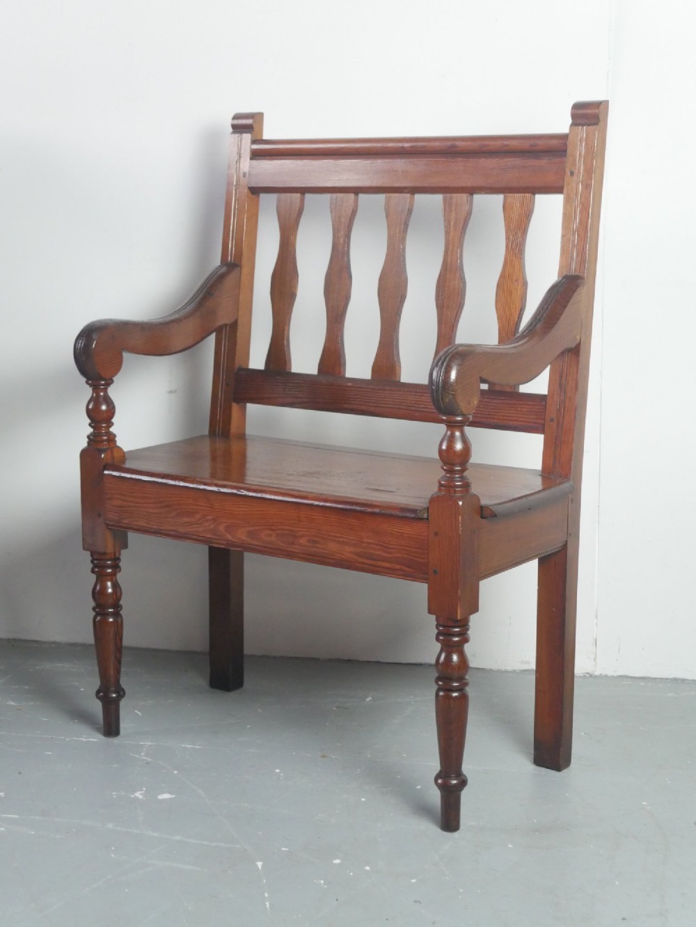 A warm brown pine hall bench featuring turned front legs, shaped back slats, swept armrests, and a solid timber seat.