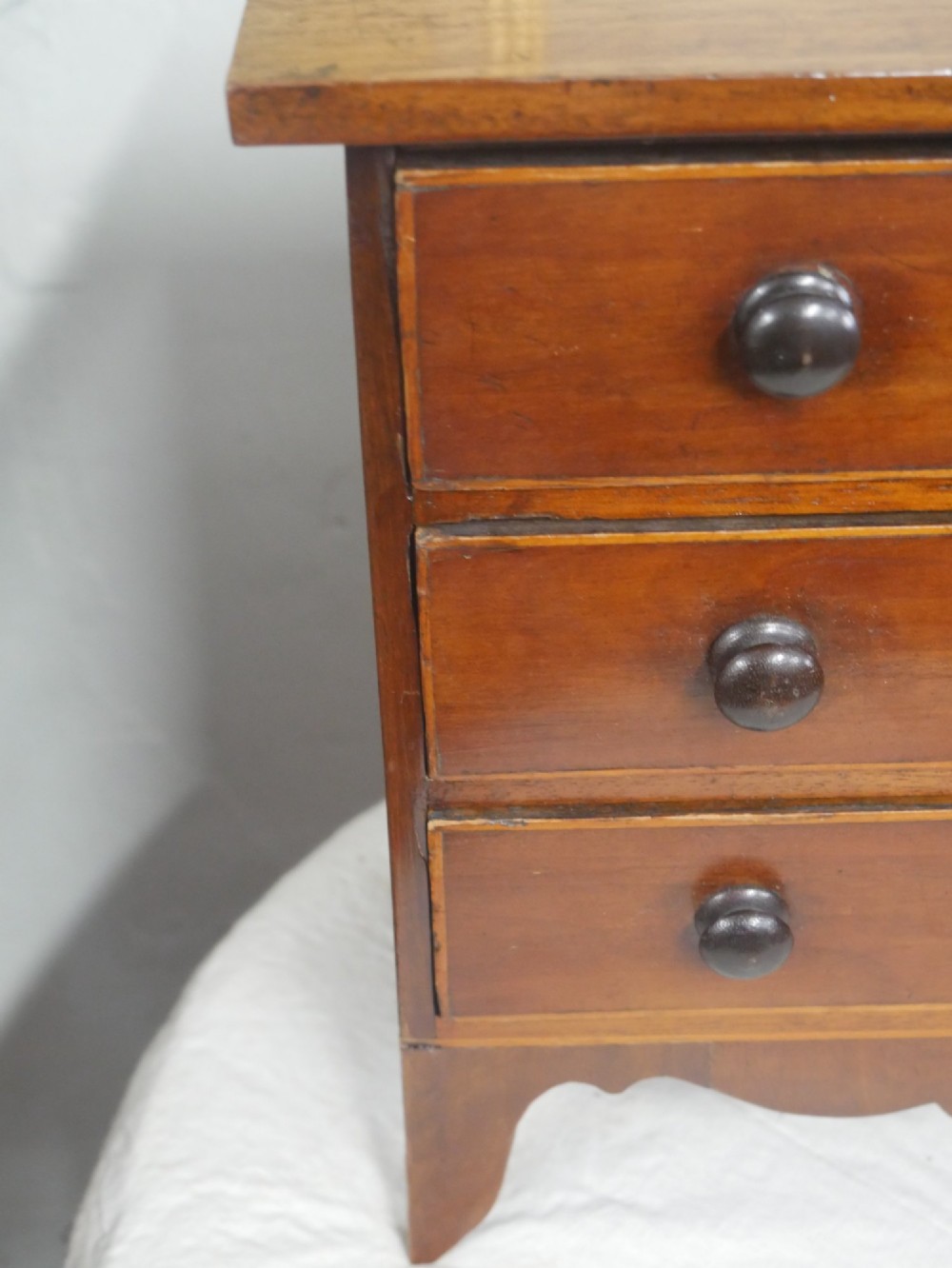 A miniature warm brown wooden chest of drawers featuring three graduated drawers with round knobs and a scalloped lower apron. View III