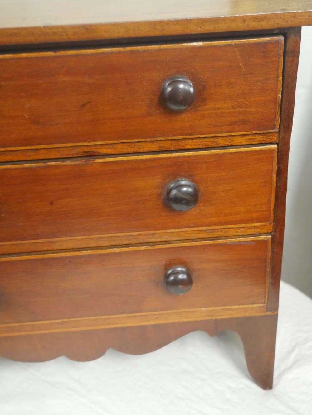 A miniature warm brown wooden chest of drawers featuring three graduated drawers with round knobs and a scalloped lower apron. View IV