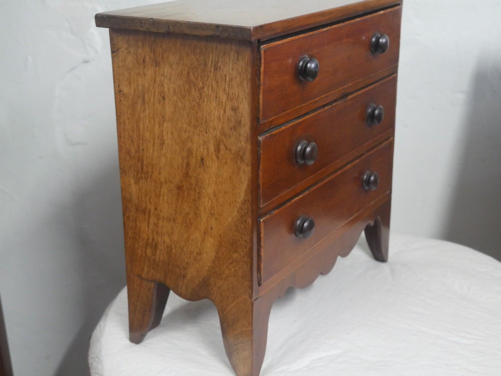 A miniature warm brown wooden chest of drawers featuring three graduated drawers with round knobs and a scalloped lower apron. View V