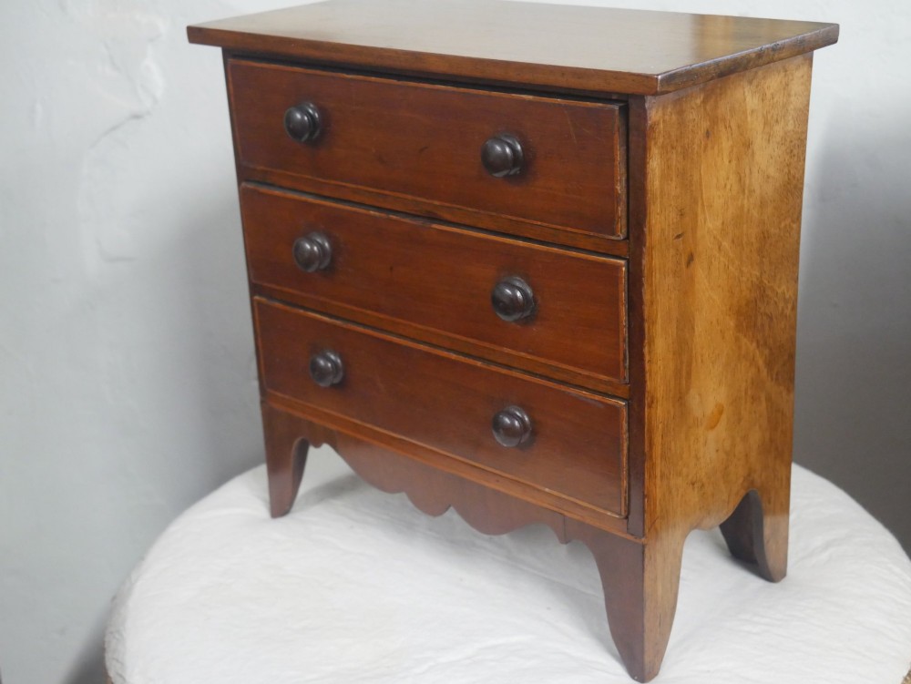 A miniature warm brown wooden chest of drawers featuring three graduated drawers with round knobs and a scalloped lower apron. View VI