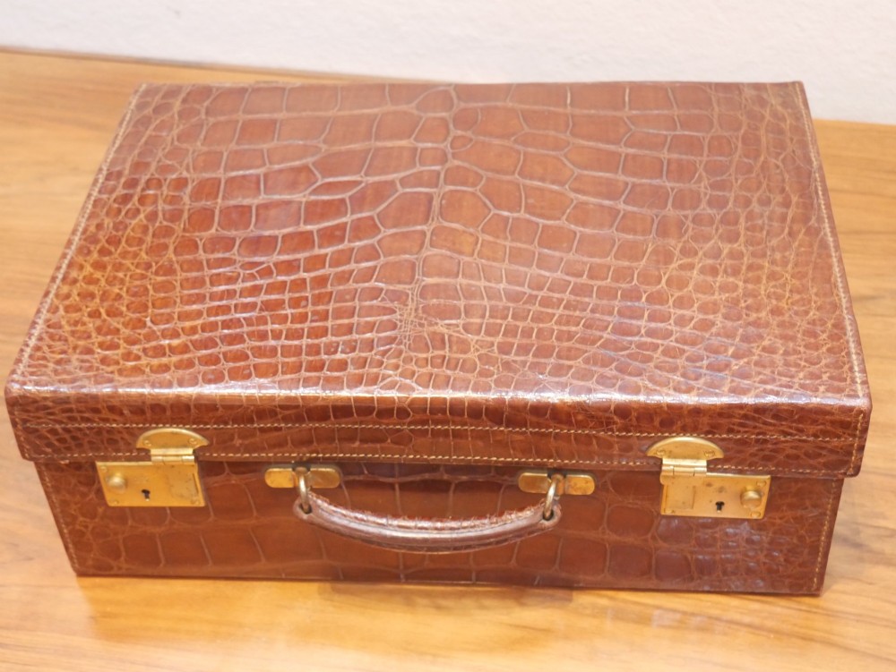 A small warm brown crocodile skin suitcase with brass locks and a stitched leather handle.