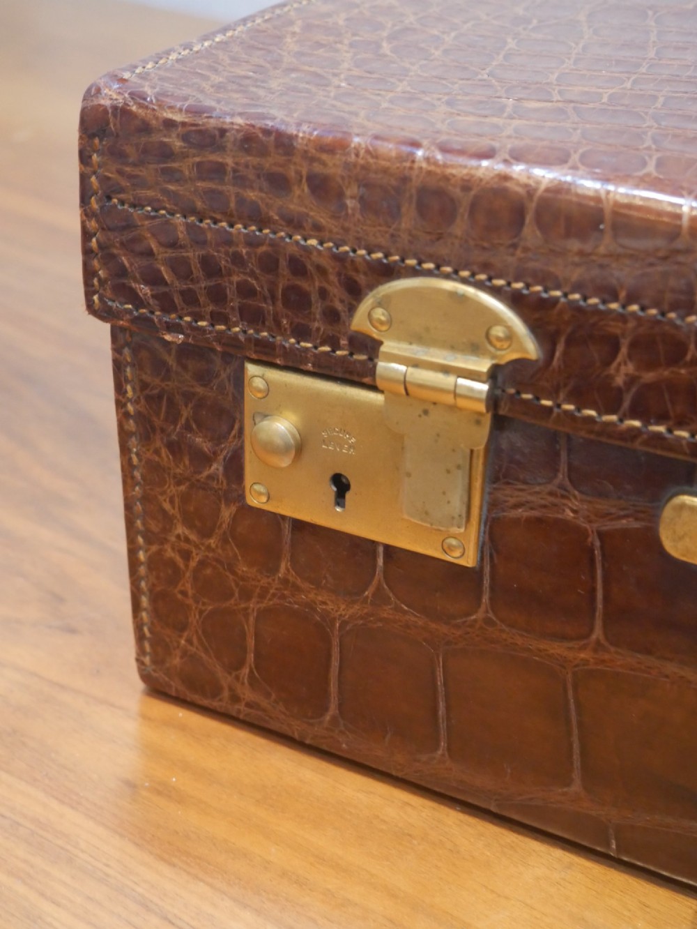 A small warm brown crocodile skin suitcase with brass locks and a stitched leather handle. View II