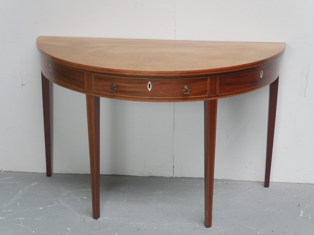 A Regency mahogany demi-lune console table featuring satinwood stringing inlay, a central drawer, and four square tapered legs.