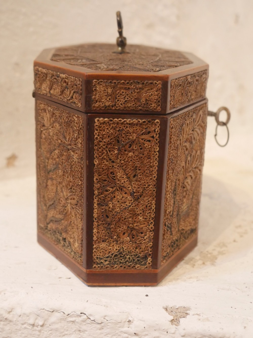 An 18th-century octagonal wooden tea caddy featuring intricate paper filigree quilling, foliate designs, a top brass loop handle, and a key. View VII