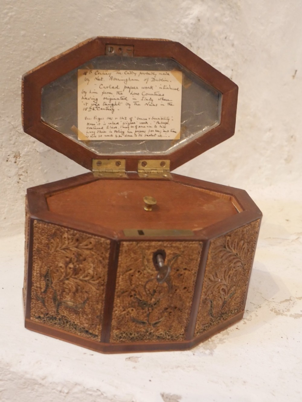 An 18th-century octagonal wooden tea caddy featuring intricate paper filigree quilling, foliate designs, a top brass loop handle, and a key. View X