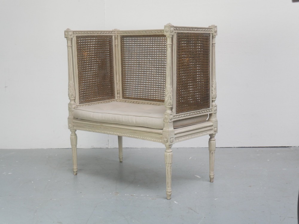 A painted off-white neoclassical window seat featuring high woven cane side and back panels, turned fluted legs, and a fitted cream cushion. View II