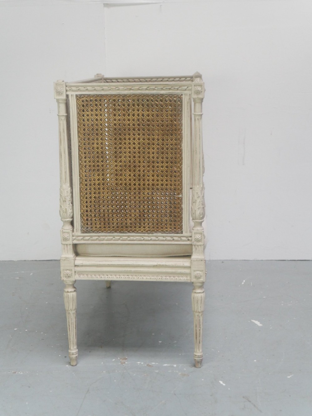 A painted off-white neoclassical window seat featuring high woven cane side and back panels, turned fluted legs, and a fitted cream cushion. View IV