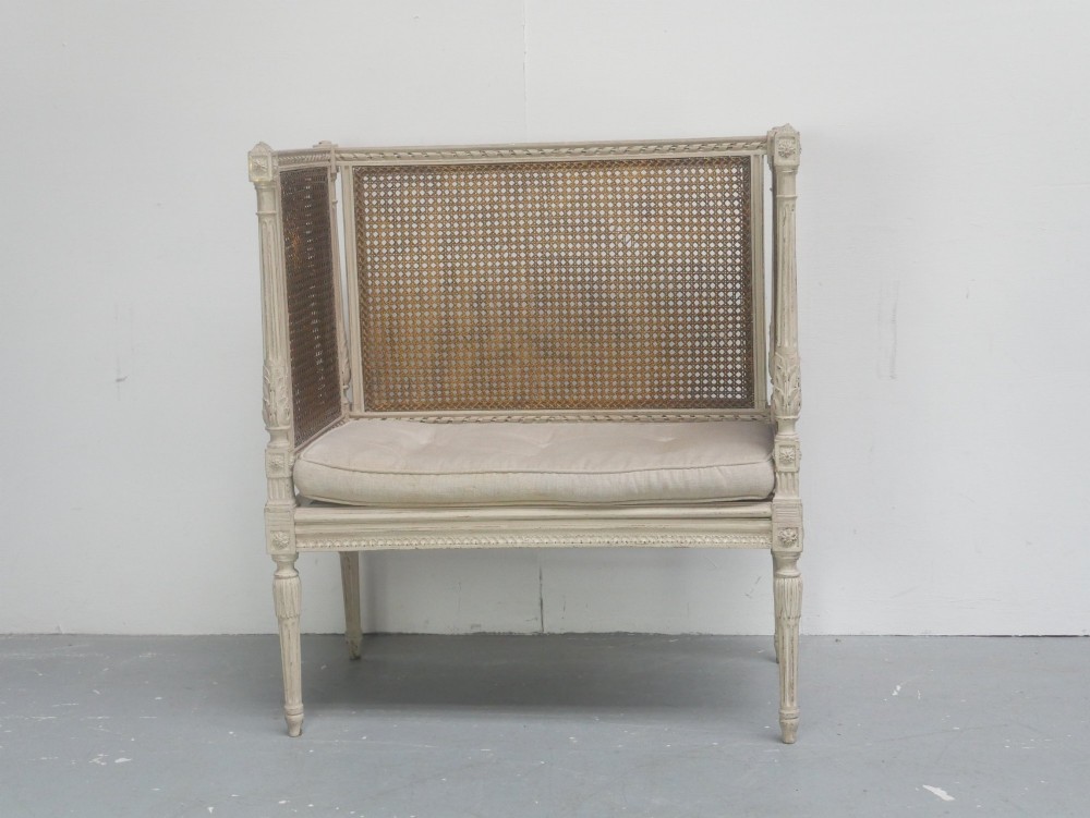 A painted off-white neoclassical window seat featuring high woven cane side and back panels, turned fluted legs, and a fitted cream cushion.