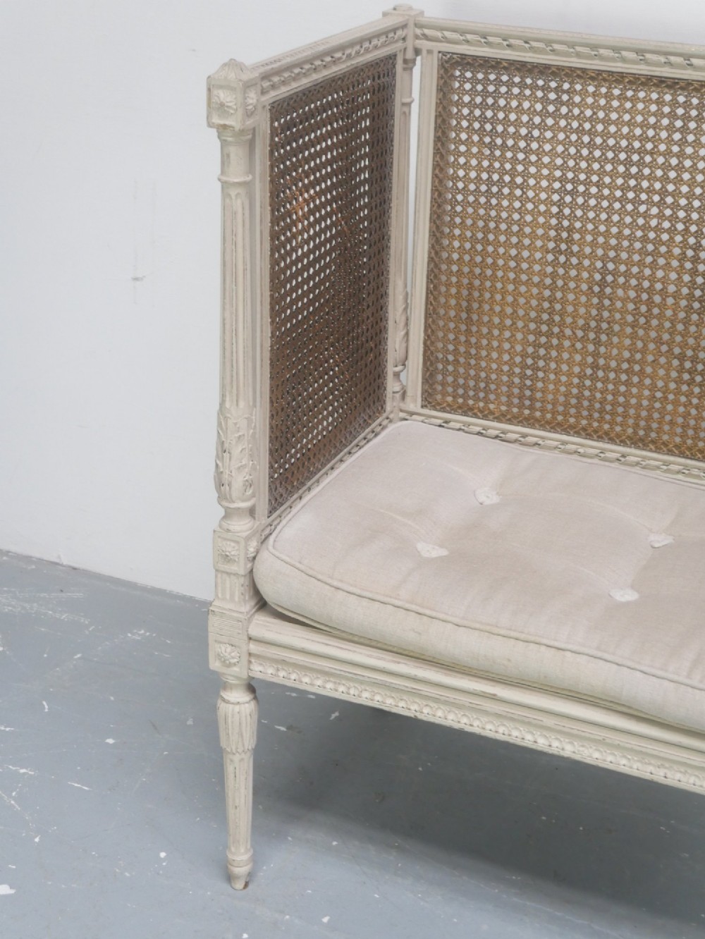 A painted off-white neoclassical window seat featuring high woven cane side and back panels, turned fluted legs, and a fitted cream cushion. View VI