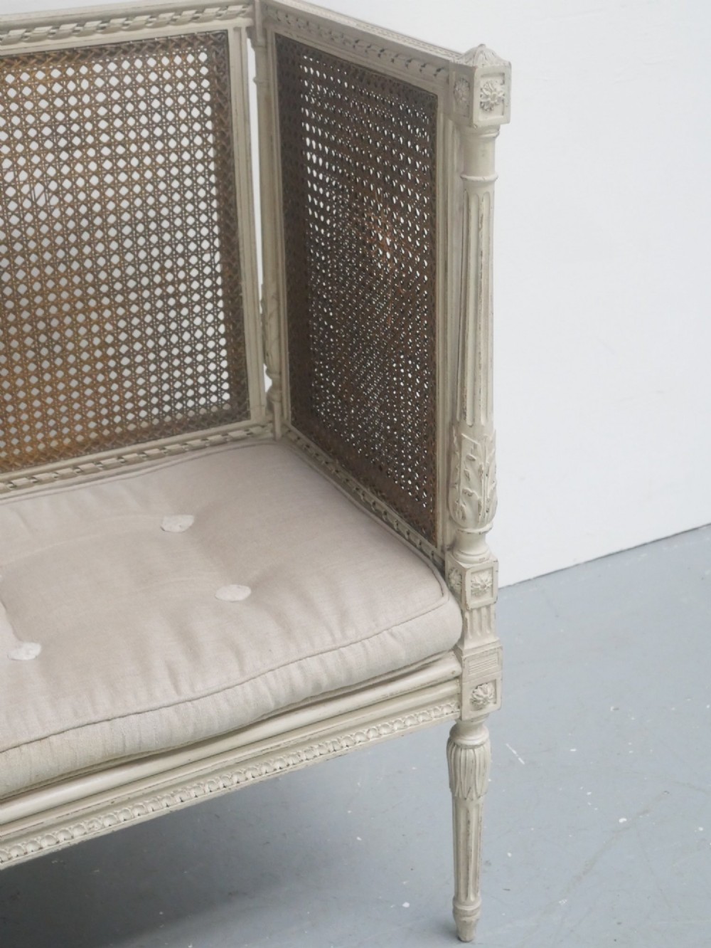 A painted off-white neoclassical window seat featuring high woven cane side and back panels, turned fluted legs, and a fitted cream cushion. View VII