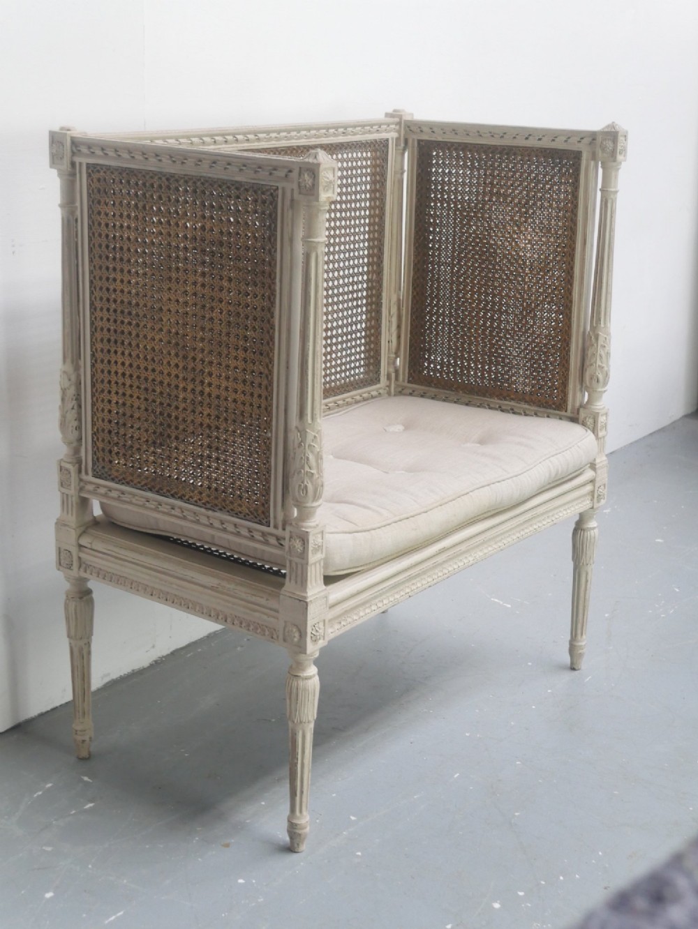 A painted off-white neoclassical window seat featuring high woven cane side and back panels, turned fluted legs, and a fitted cream cushion. View IX