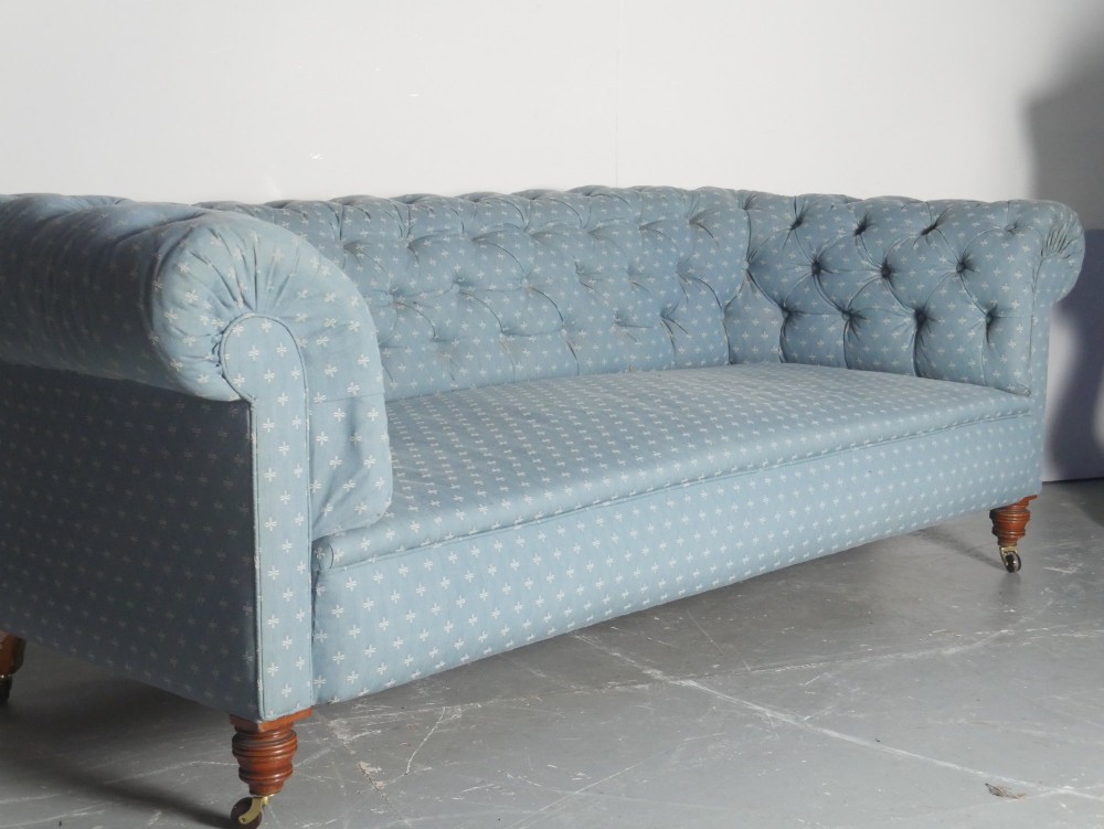 A light blue patterned fabric Chesterfield sofa featuring deep buttoning across the backrest and rolled arms, supported by turned wooden legs on brass castors.