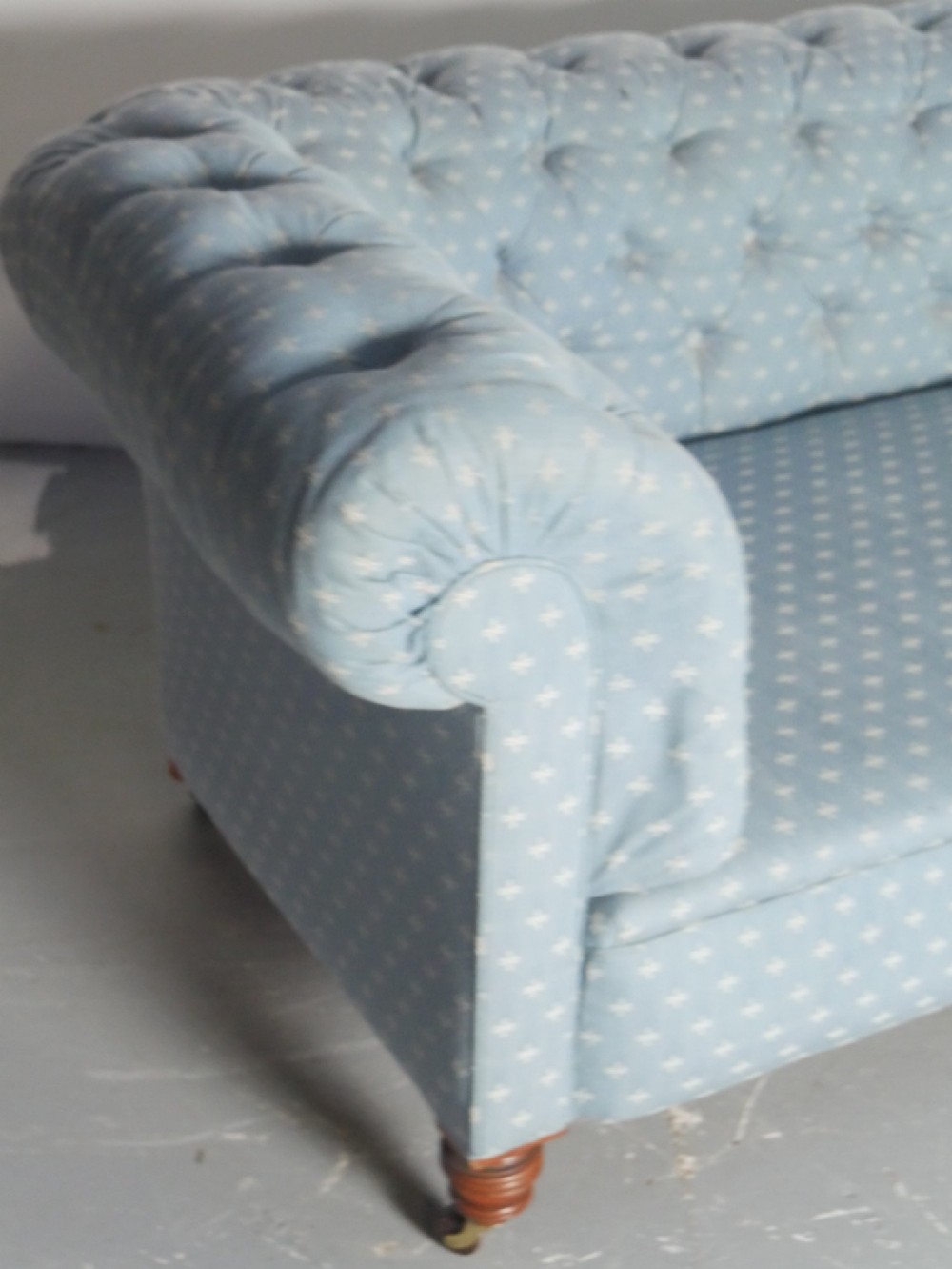 A light blue patterned fabric Chesterfield sofa featuring deep buttoning across the backrest and rolled arms, supported by turned wooden legs on brass castors. View II