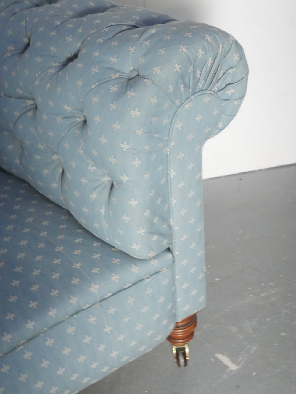 A light blue patterned fabric Chesterfield sofa featuring deep buttoning across the backrest and rolled arms, supported by turned wooden legs on brass castors. View IV