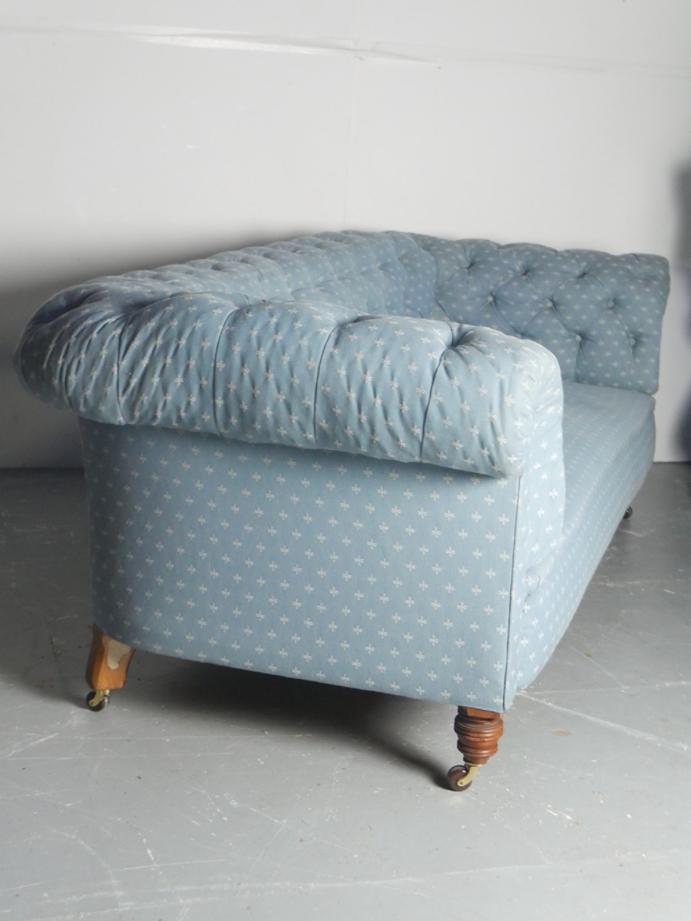 A light blue patterned fabric Chesterfield sofa featuring deep buttoning across the backrest and rolled arms, supported by turned wooden legs on brass castors. View V
