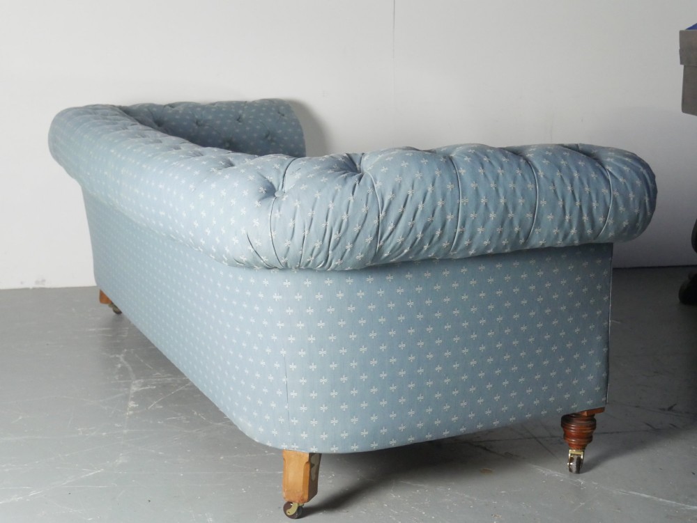 A light blue patterned fabric Chesterfield sofa featuring deep buttoning across the backrest and rolled arms, supported by turned wooden legs on brass castors. View VI