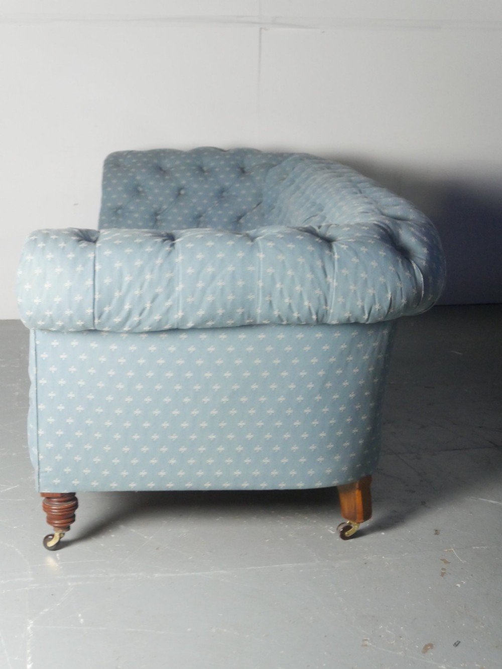 A light blue patterned fabric Chesterfield sofa featuring deep buttoning across the backrest and rolled arms, supported by turned wooden legs on brass castors. View VII