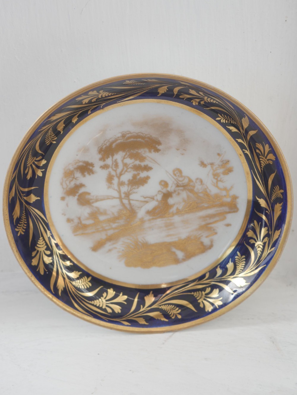 An antique porcelain teacup and matching saucer featuring cobalt blue borders with gold foliate motifs and central monochrome pastoral scenes. View II