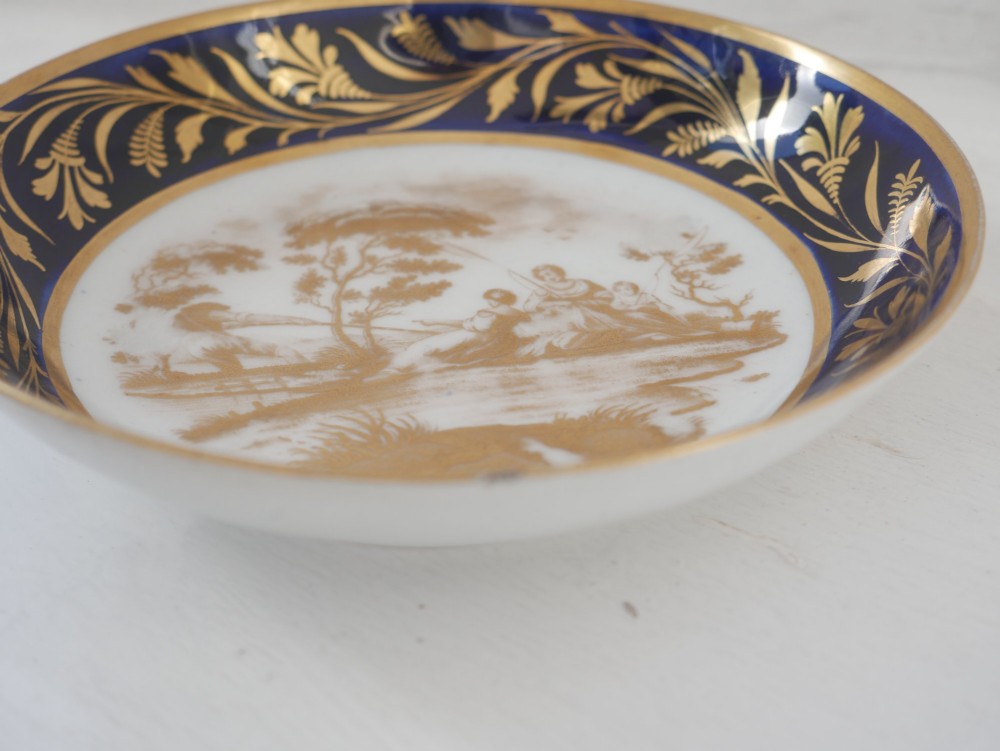 An antique porcelain teacup and matching saucer featuring cobalt blue borders with gold foliate motifs and central monochrome pastoral scenes. View III