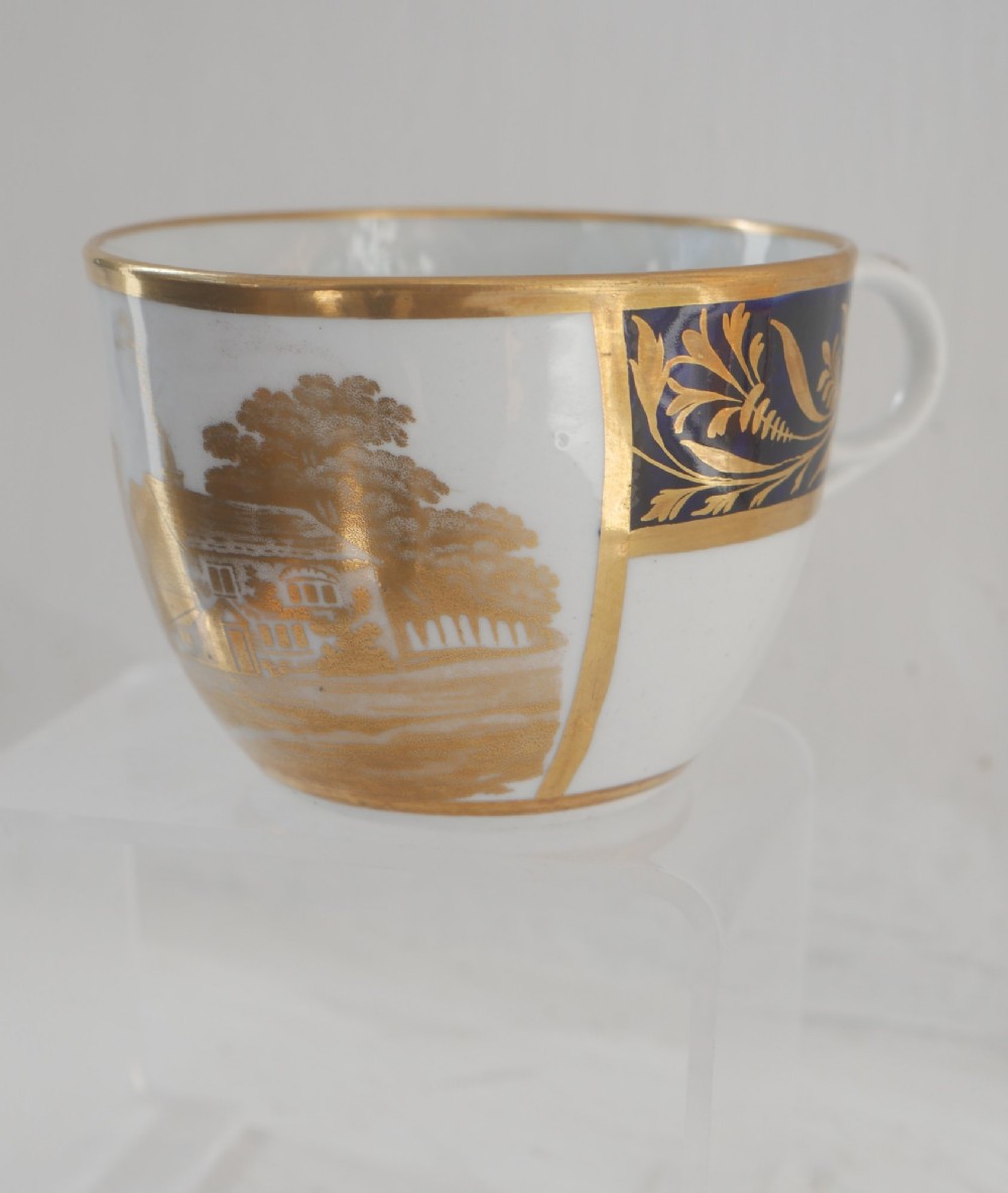 An antique porcelain teacup and matching saucer featuring cobalt blue borders with gold foliate motifs and central monochrome pastoral scenes. View V