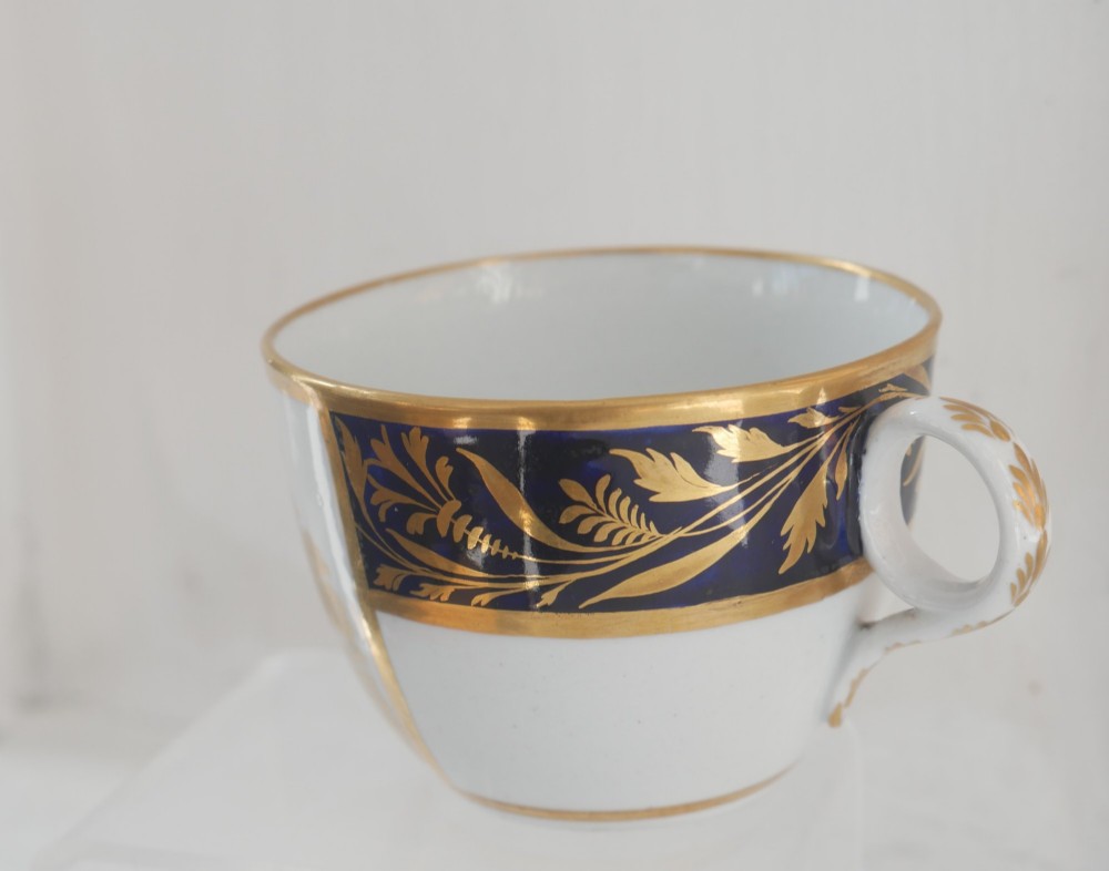 An antique porcelain teacup and matching saucer featuring cobalt blue borders with gold foliate motifs and central monochrome pastoral scenes. View VI