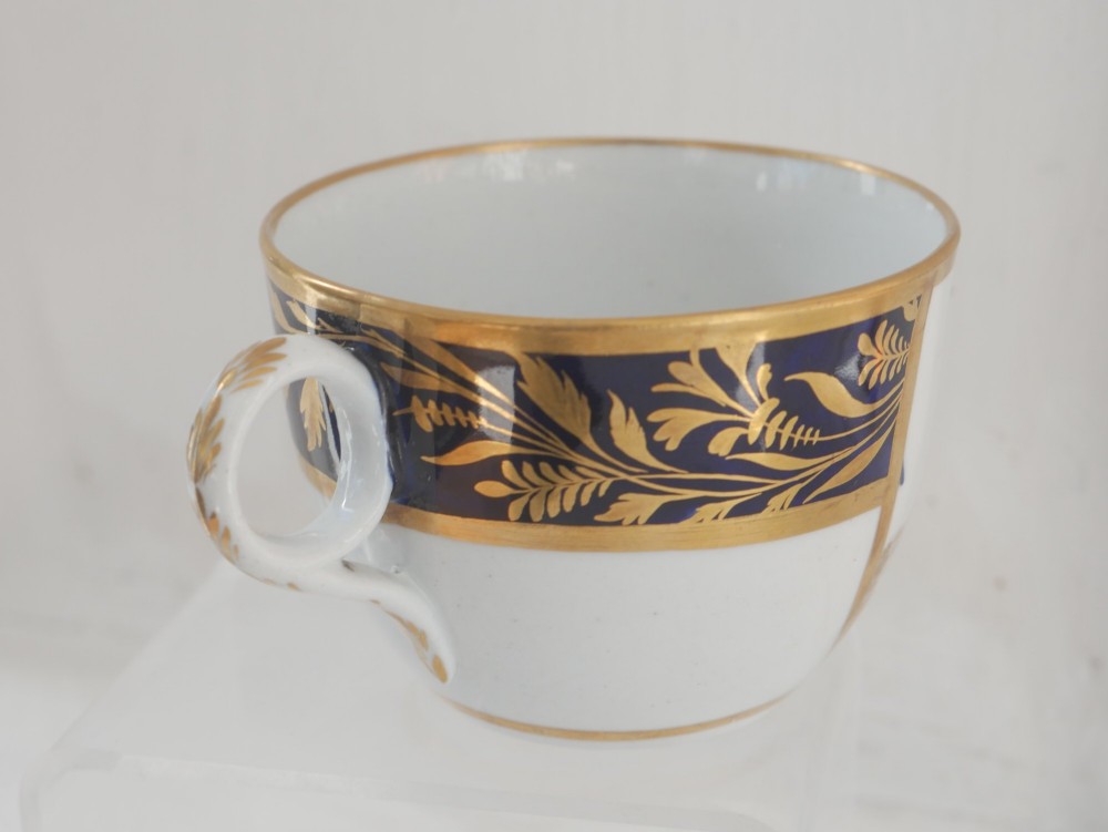 An antique porcelain teacup and matching saucer featuring cobalt blue borders with gold foliate motifs and central monochrome pastoral scenes. View VII