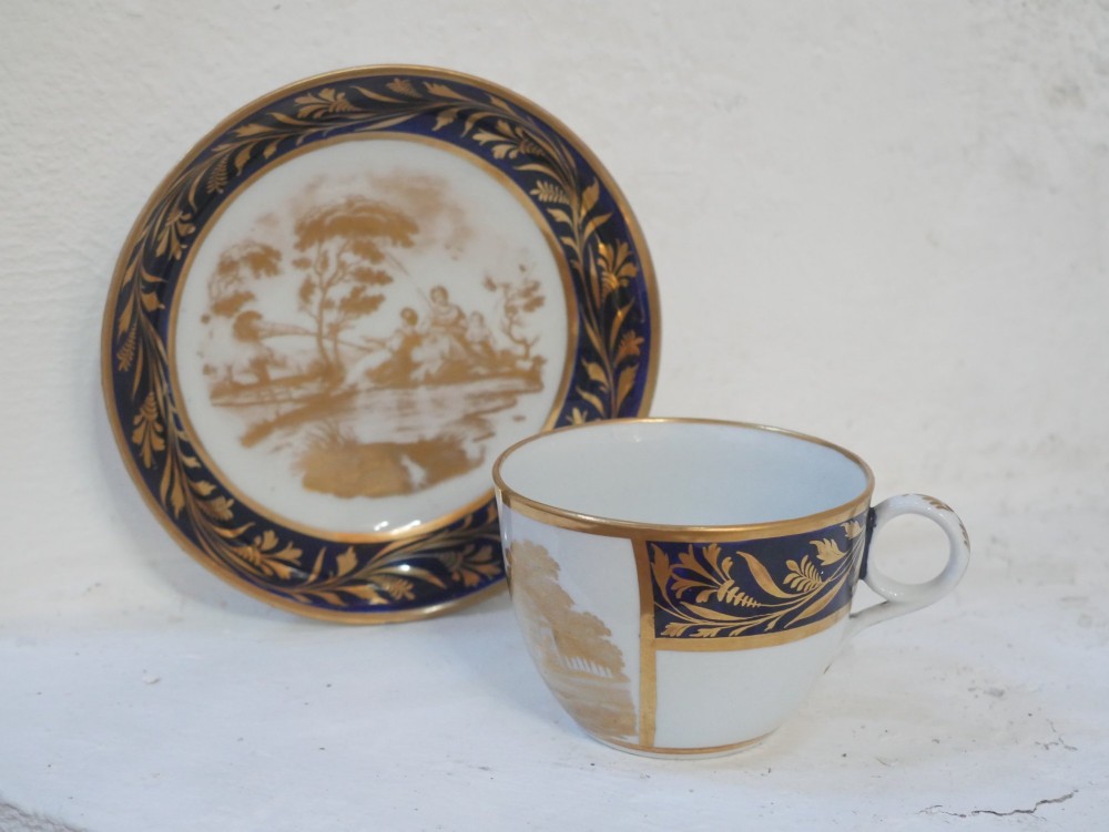 An antique porcelain teacup and matching saucer featuring cobalt blue borders with gold foliate motifs and central monochrome pastoral scenes. View IX