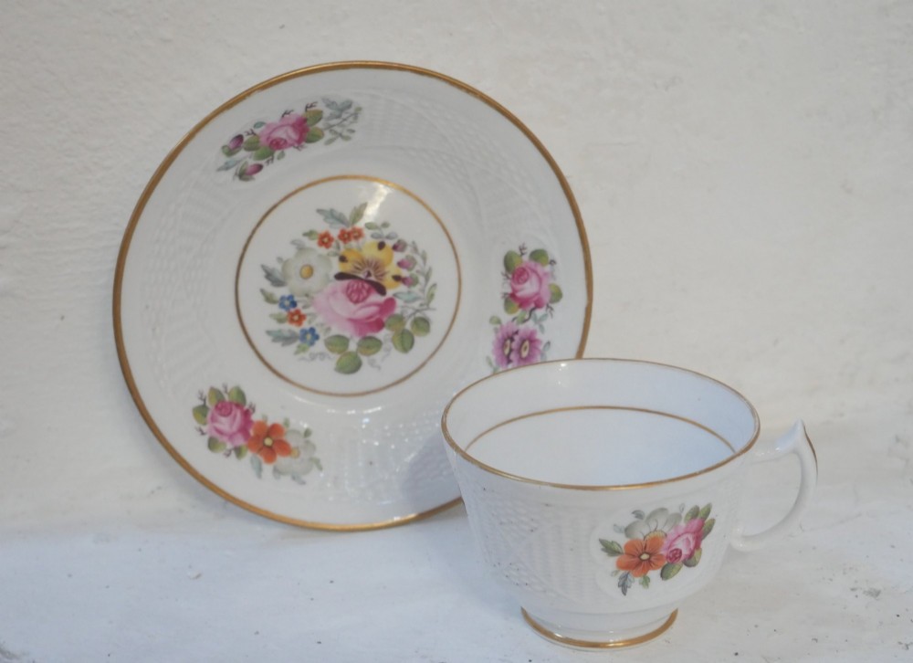 An antique white porcelain teacup and matching saucer featuring hand-painted floral sprays, moulded textured borders, and gilded rim details.
