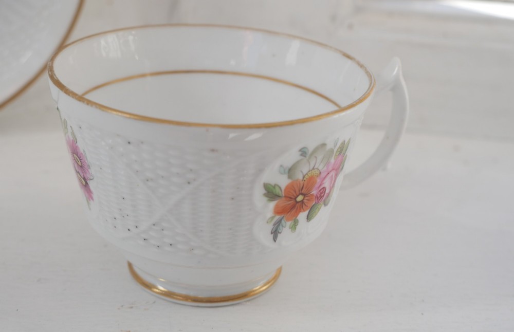 An antique white porcelain teacup and matching saucer featuring hand-painted floral sprays, moulded textured borders, and gilded rim details. View II