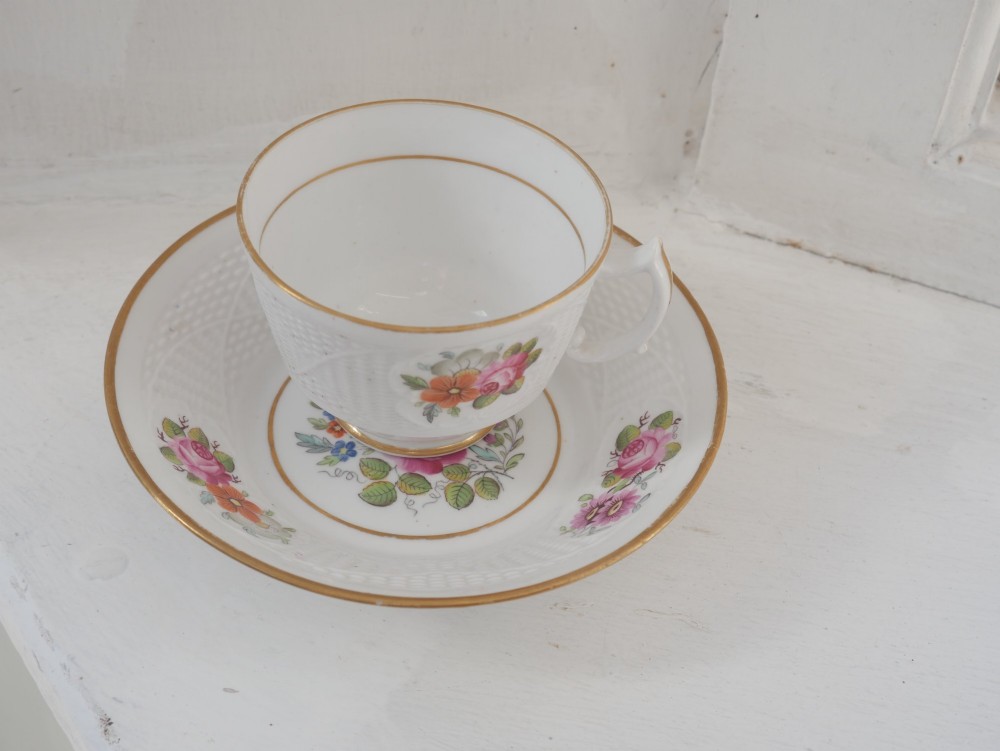 An antique white porcelain teacup and matching saucer featuring hand-painted floral sprays, moulded textured borders, and gilded rim details. View IV