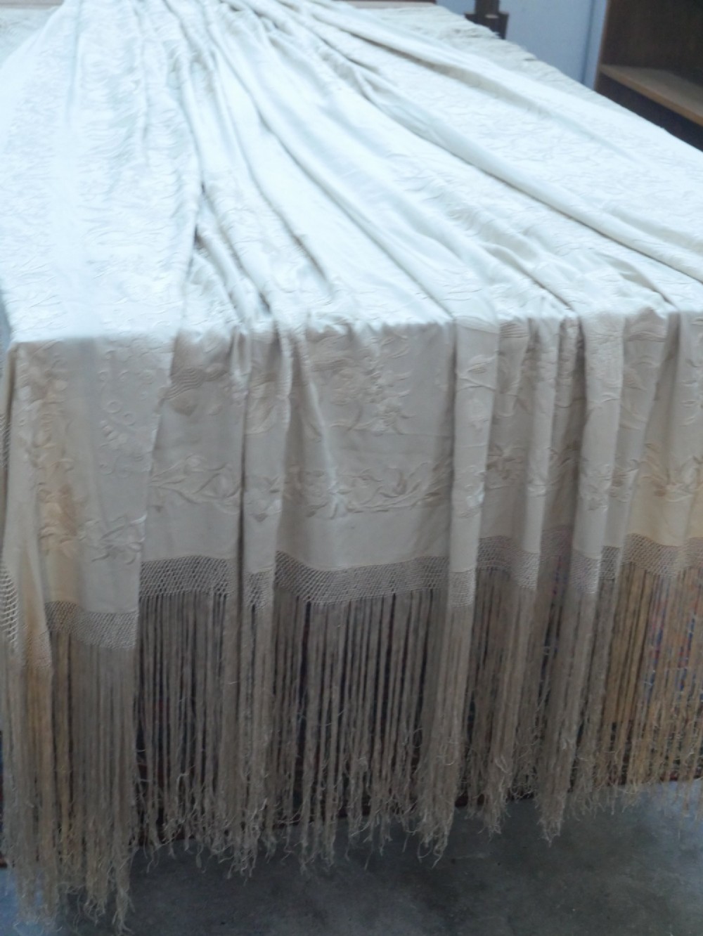 A cream-coloured silk piano shawl featuring tonal floral hand embroidery and deep knotted fringe edge.