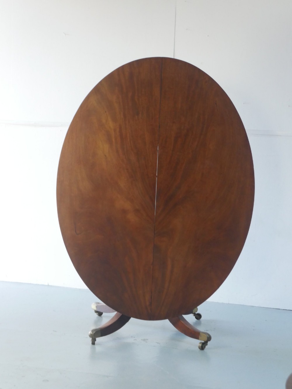 An antique Regency oval mahogany tilt-top table shown upright on a splayed pedestal base with brass castors.