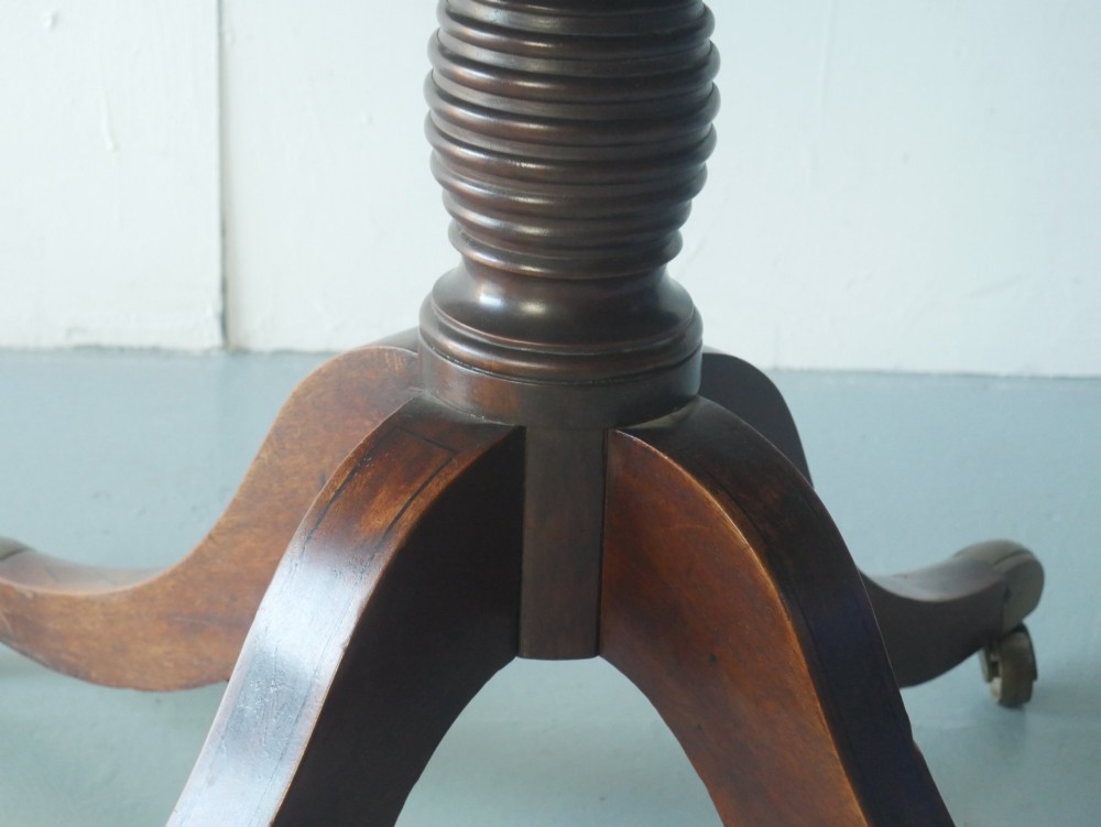 An antique Regency oval mahogany tilt-top table shown upright on a splayed pedestal base with brass castors. View III