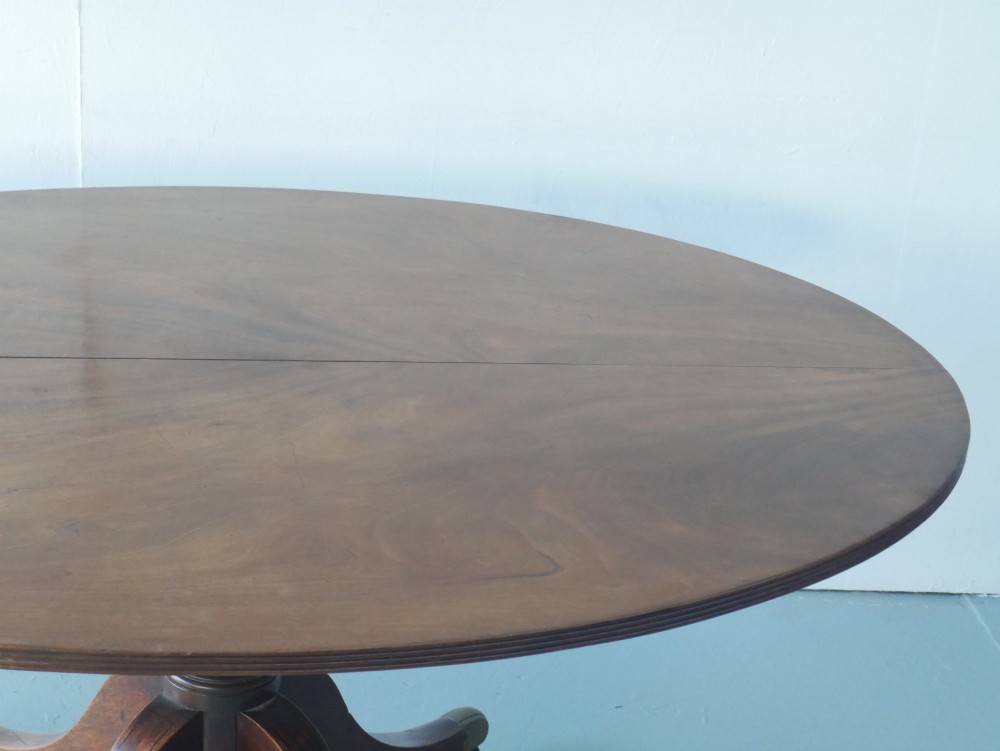 An antique Regency oval mahogany tilt-top table shown upright on a splayed pedestal base with brass castors. View IV